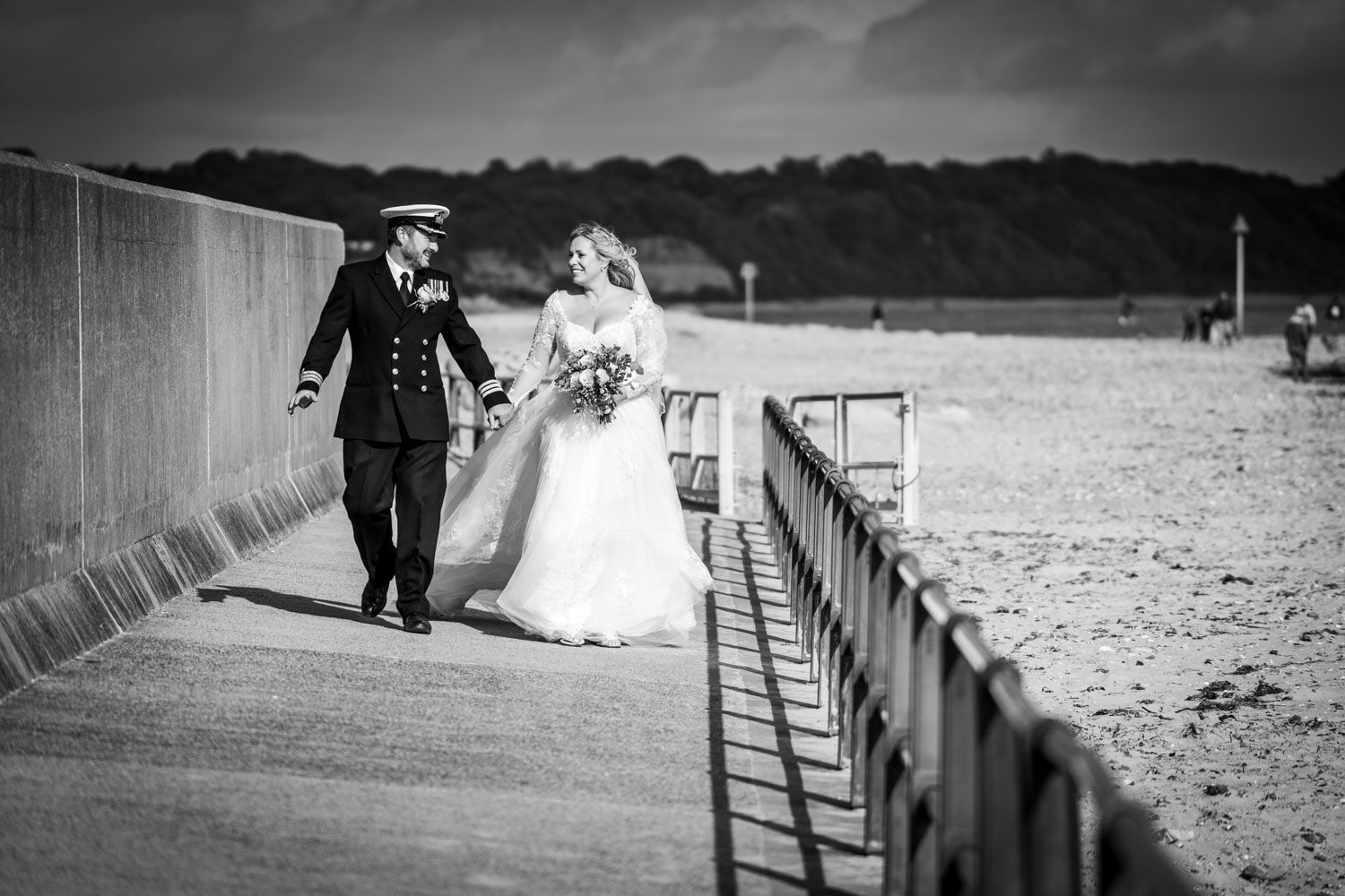 Black and white photograph of a couple in wedding attire walking along a seaside pier on their special day.