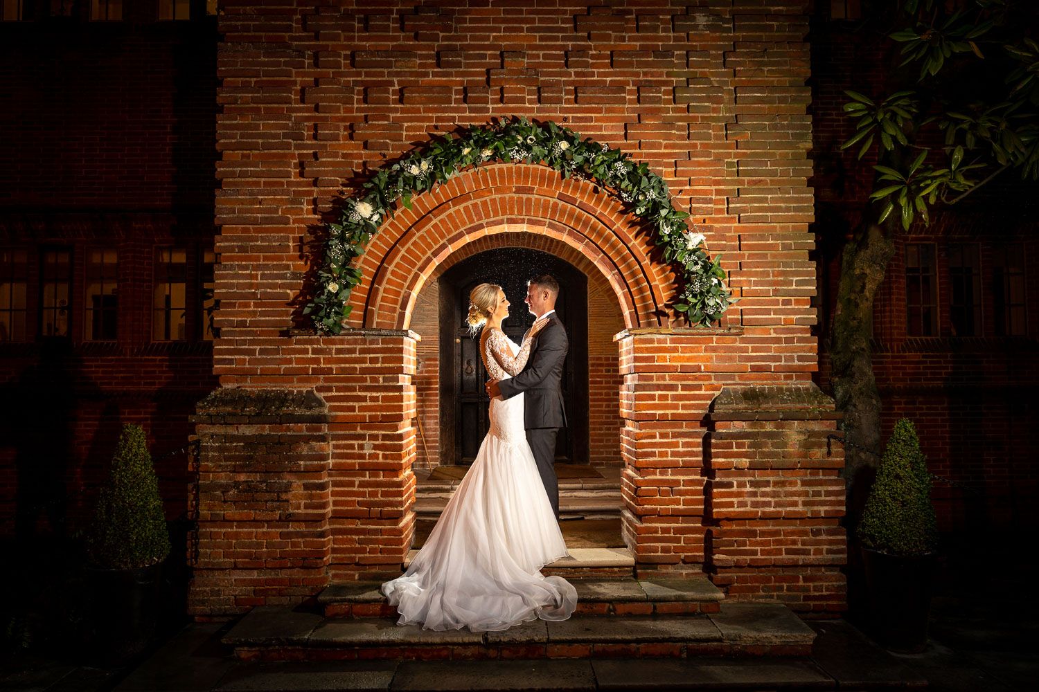 A wedding couple poses under a brick archway decorated with holiday greenery and lights.