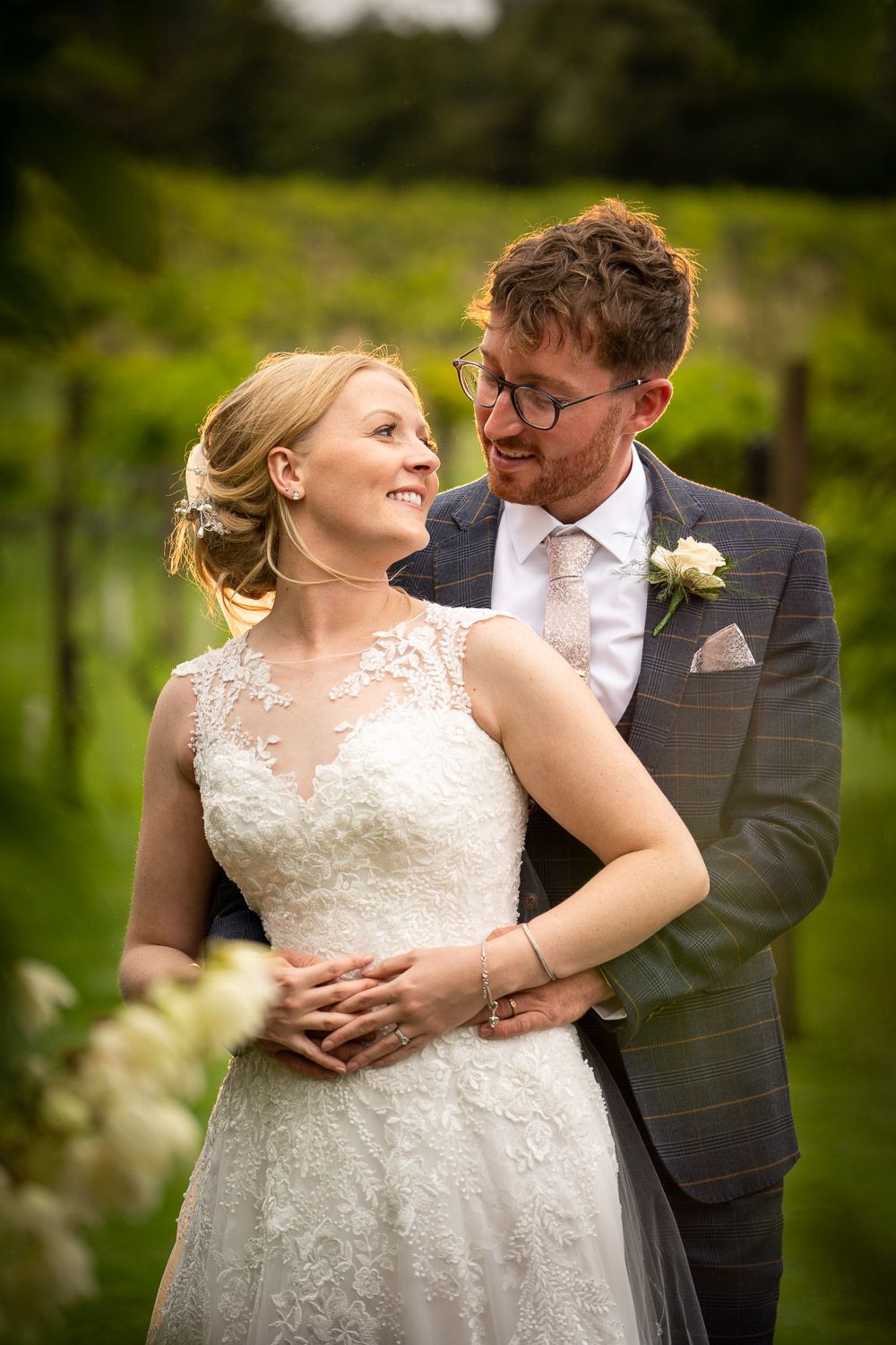 A bride in a white lace gown and groom in a dark suit share an intimate moment in a garden setting.