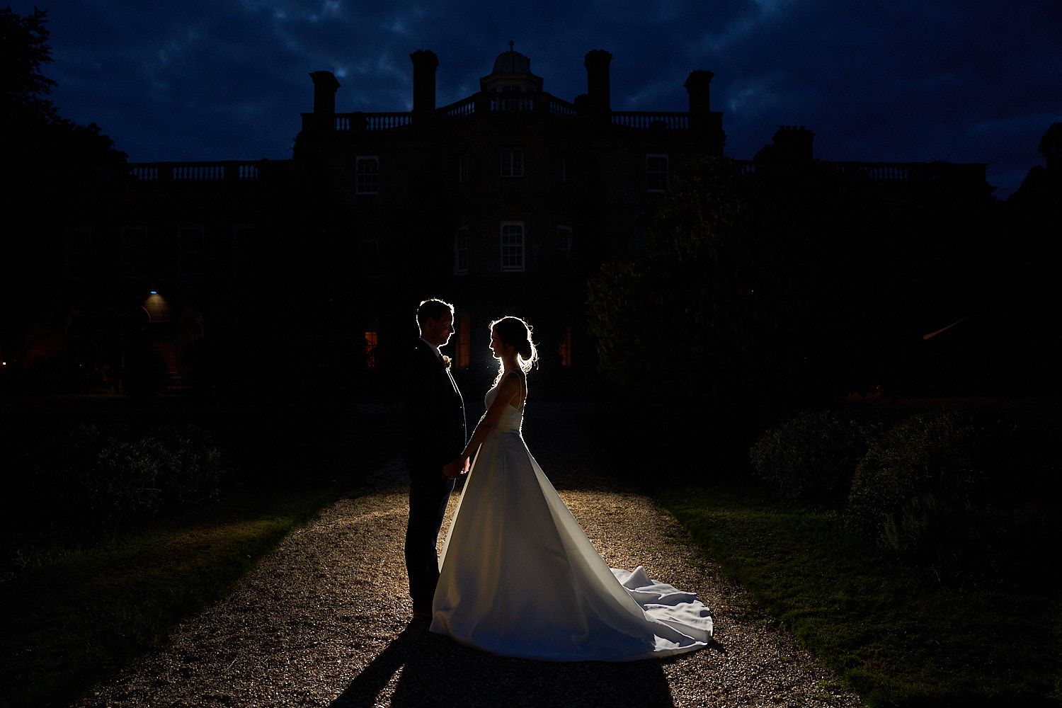 A backlit couple stands on a gravel path at dusk with a grand manor house silhouetted in the background.