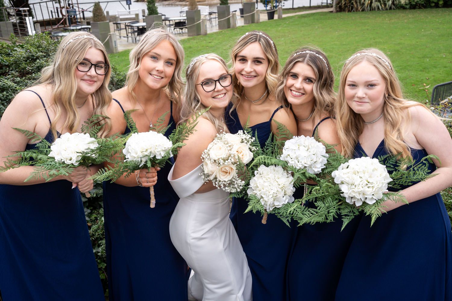 Bridesmaids in elegant navy blue dresses pose together holding white hydrangea bouquets at outdoor wedding.