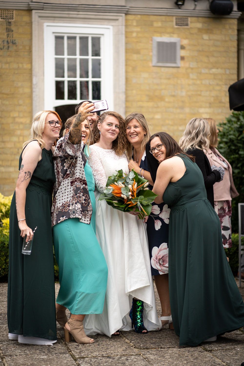 A group standing together at a wedding celebration wearing dark green dresses and floral prints around a white gown.