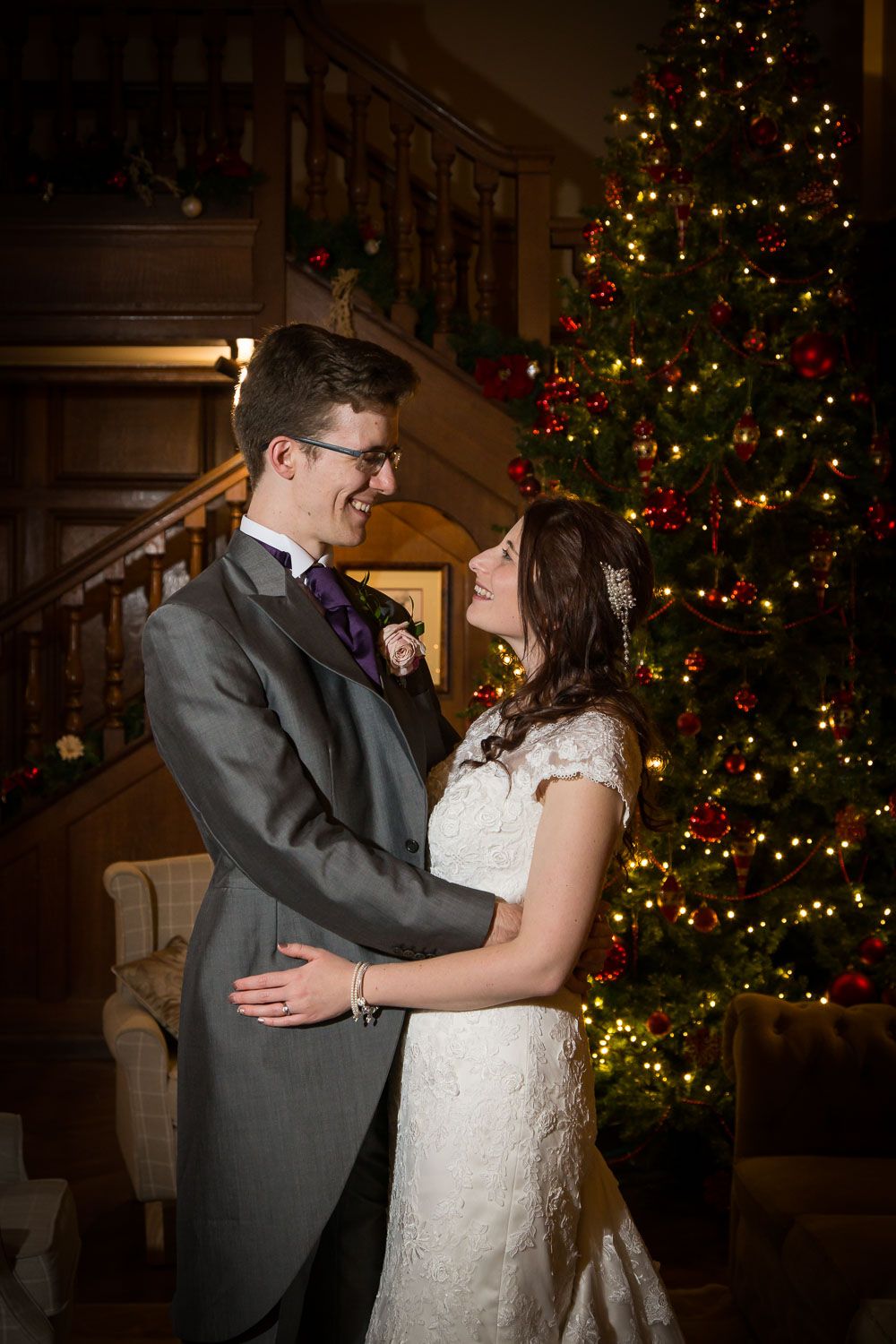 A couple in wedding attire shares a romantic moment in front of a decorated Christmas tree in a warmly lit room.