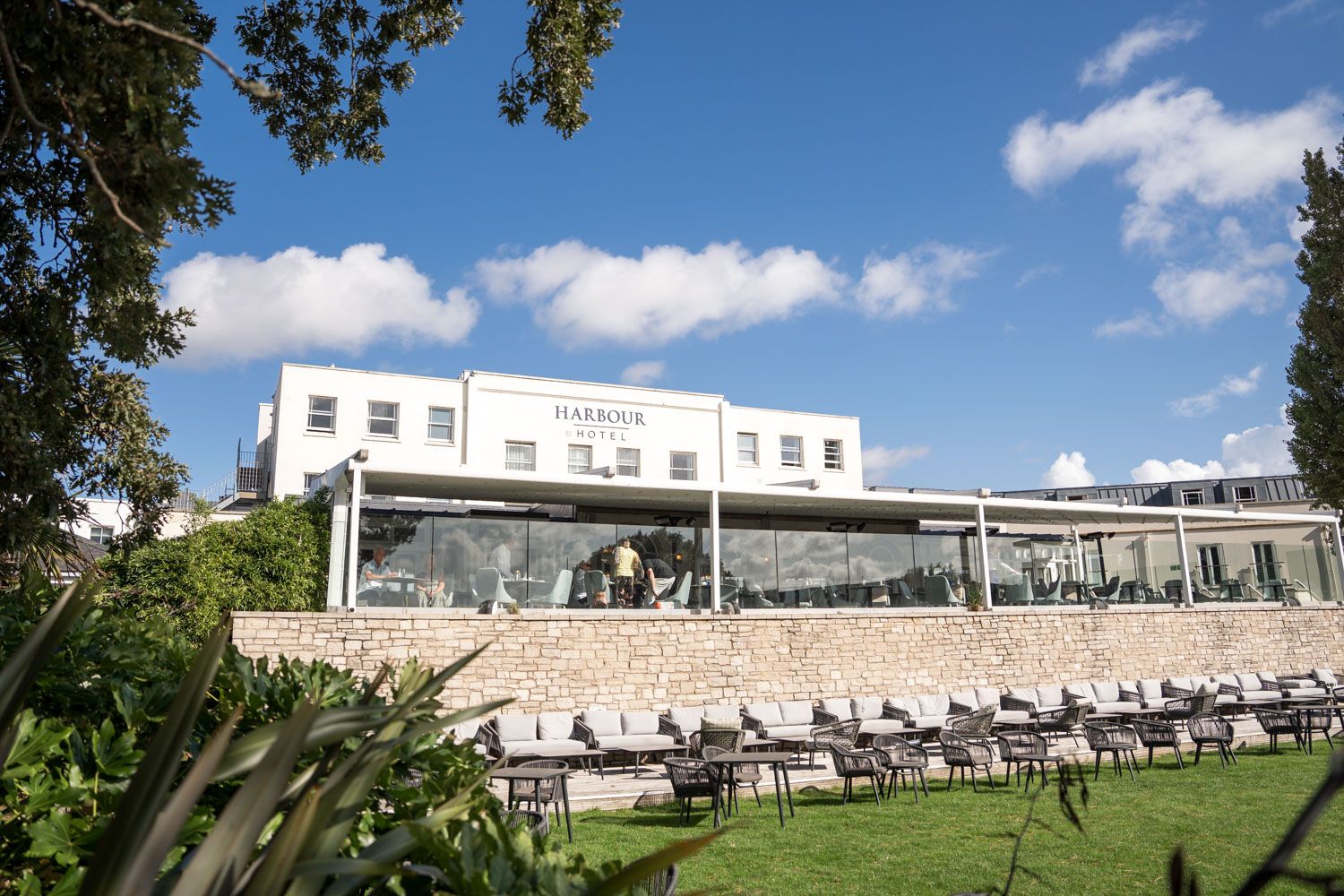 Modern building facade with outdoor seating and tables arranged under white awning by large glass windows in sunny weather.