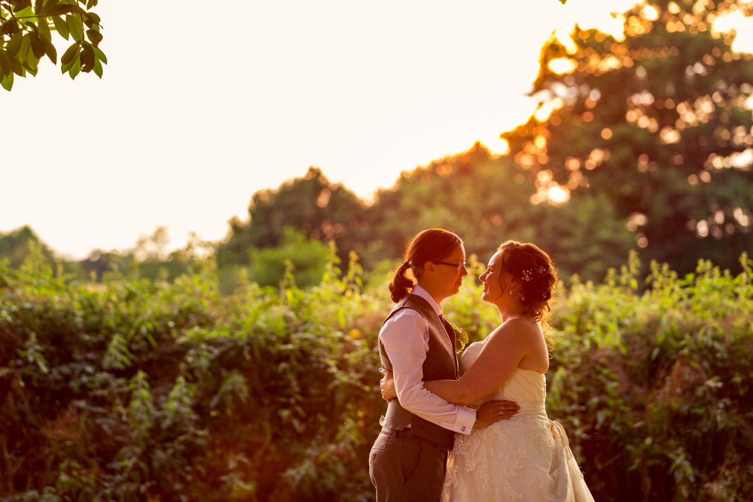 Two brides share tender moment at golden sunset in lush garden setting.
