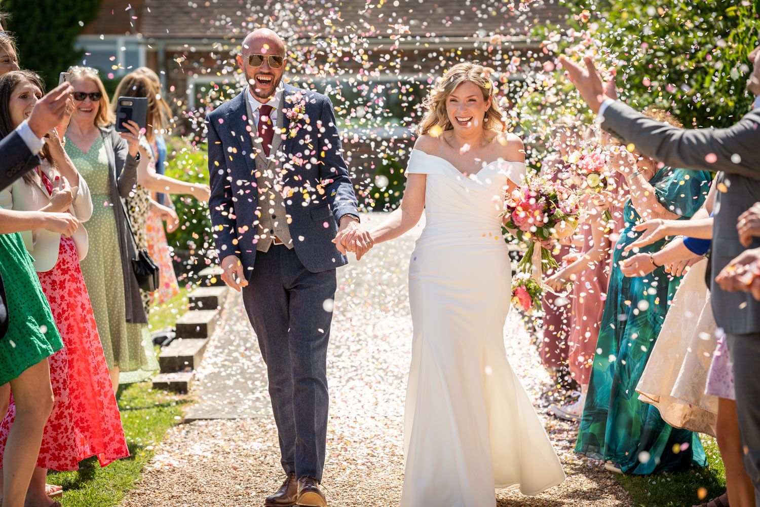 Wedding guests shower a couple with confetti as they walk down a path in celebration.
