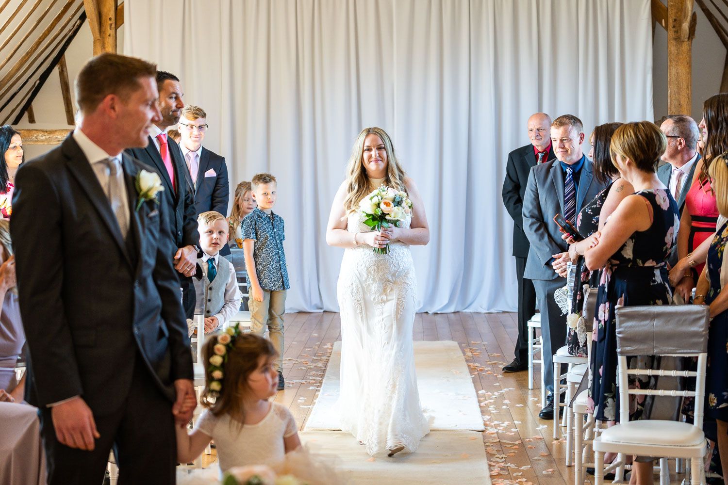 Elegant wedding ceremony in a barn venue with white draping and guests seated in wooden chairs along the aisle.