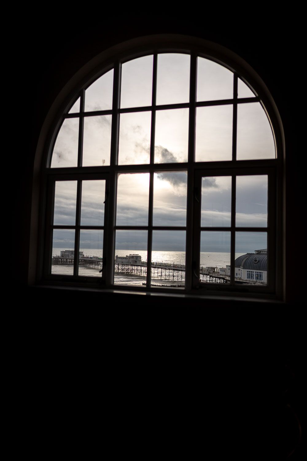 An arched window frames a moody coastal scene with dark clouds looming over a pier and waterfront structures.