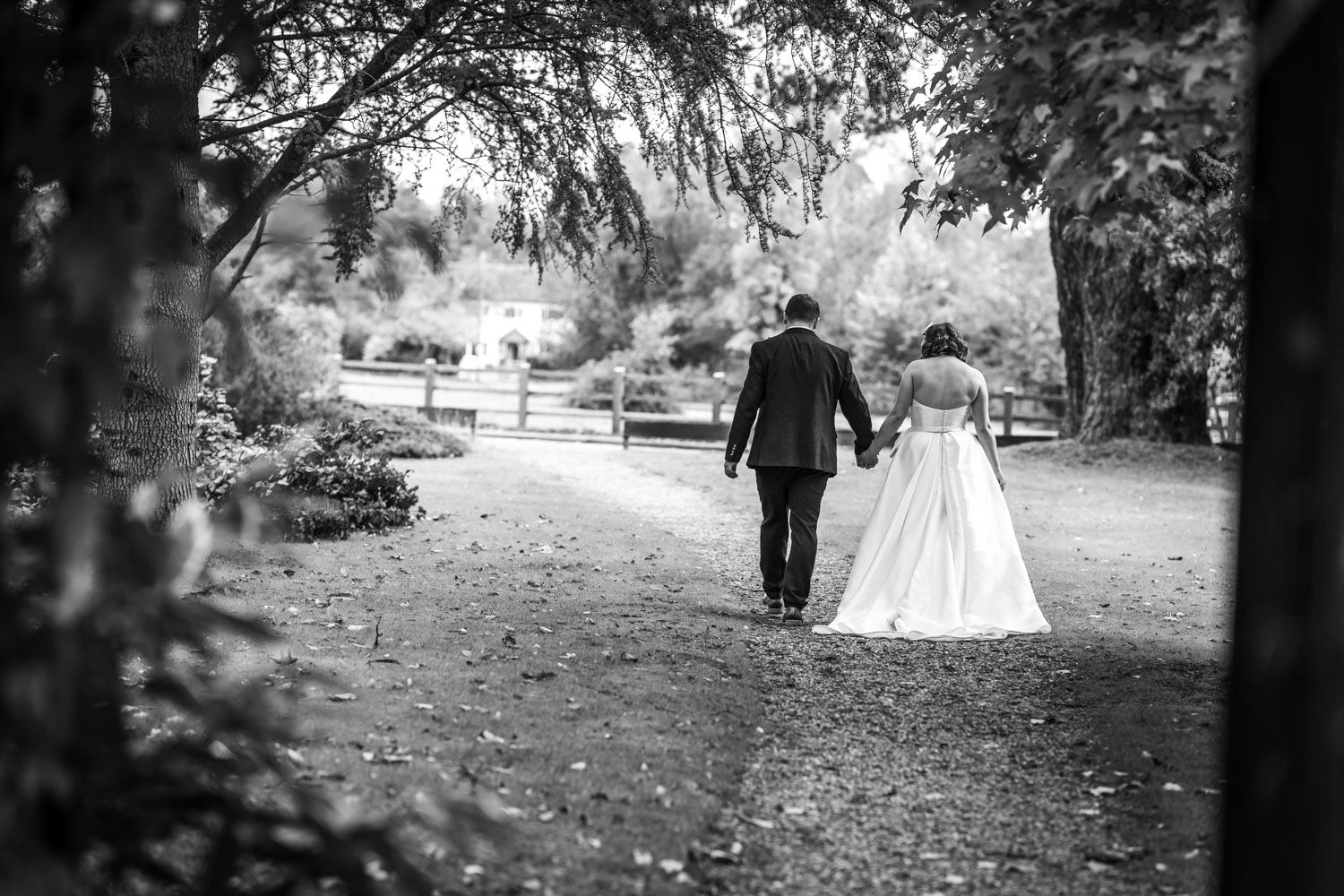 Black and white wedding photography shows couple walking down garden path under overhanging trees at romantic venue.