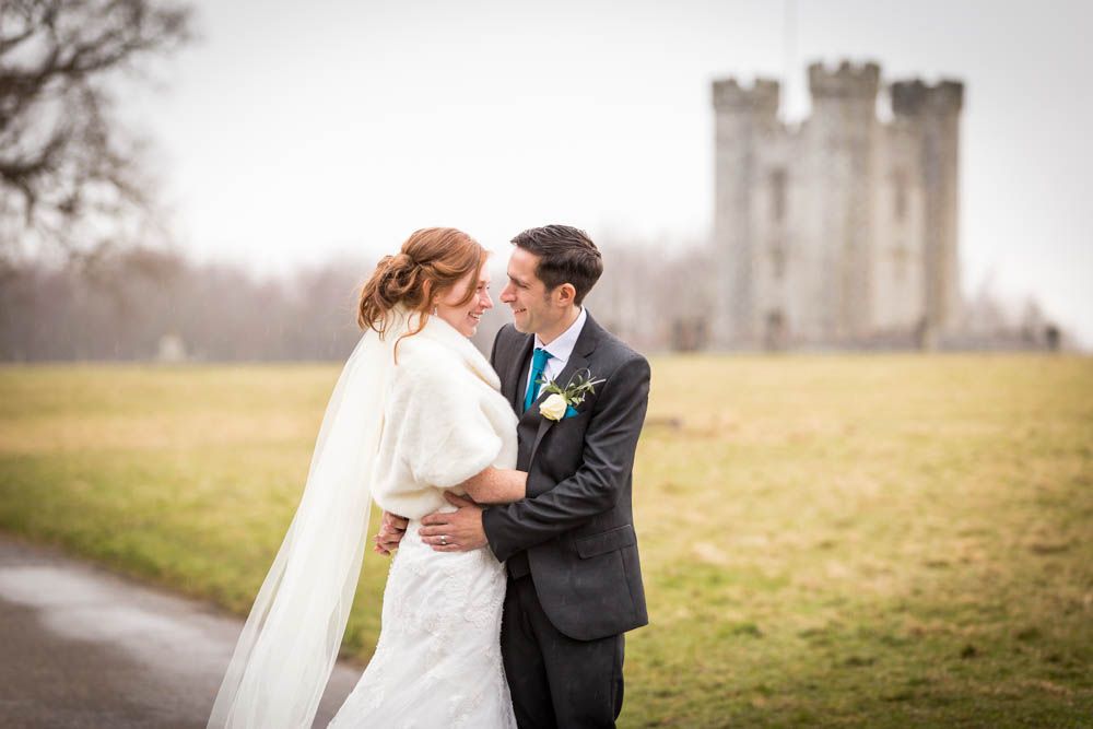 Two people in wedding attire embrace on a path with a historic stone castle tower visible in the misty background.