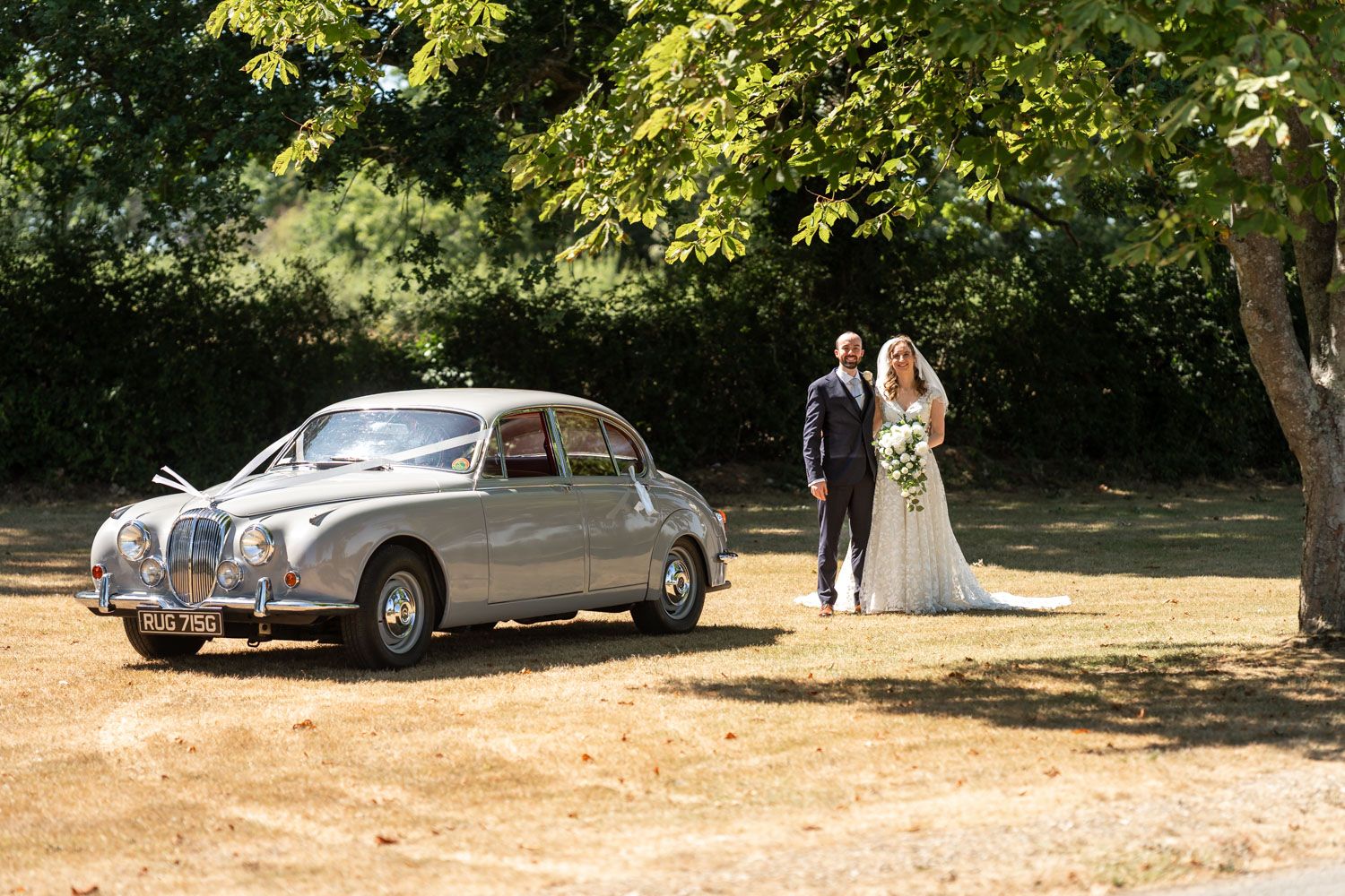 Bride and groom stand beside a classic silver vintage car under a shady tree on sunny day.