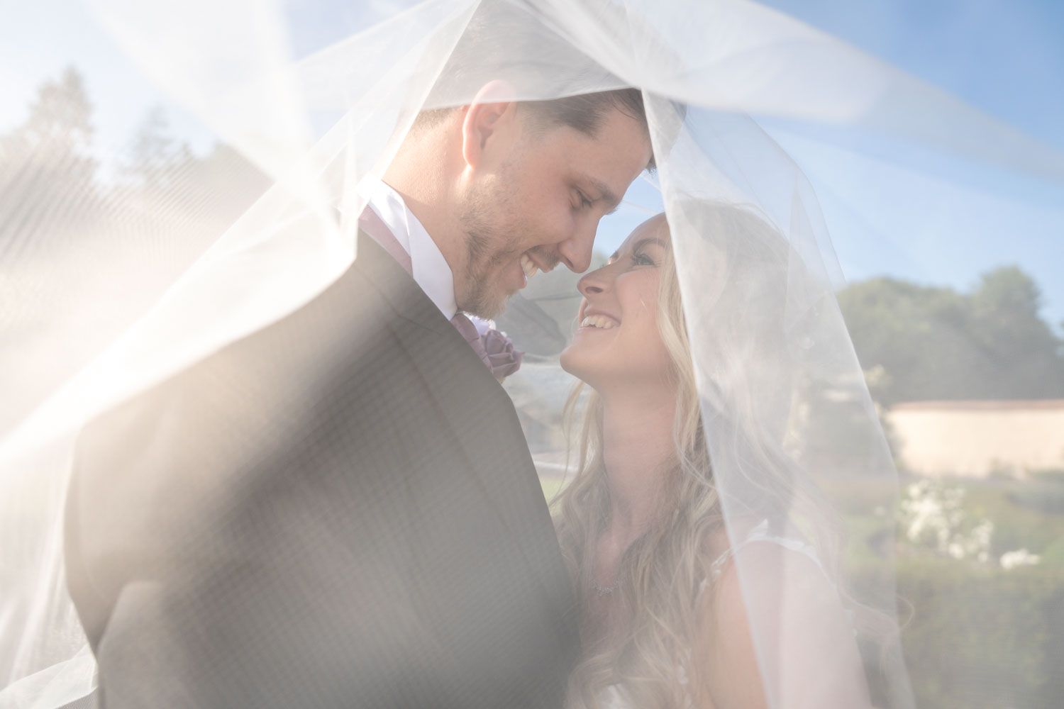 Romantic outdoor portrait of newlyweds sharing an intimate moment under a flowing veil with soft sunlight.