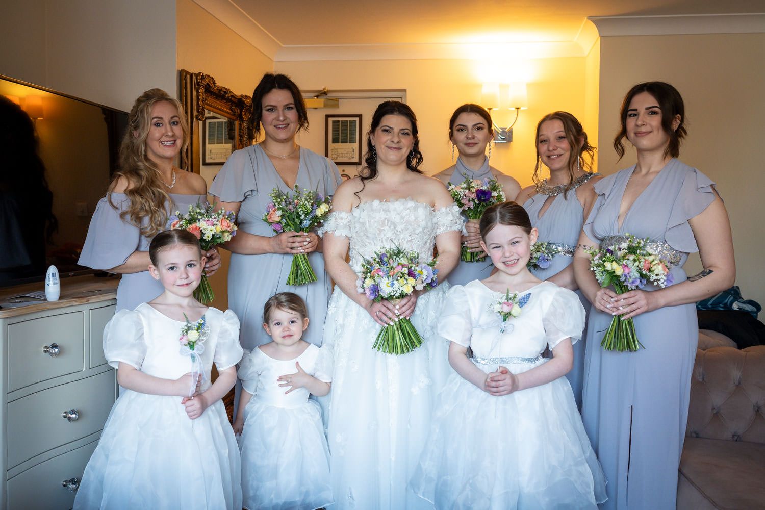 A bridal party poses together in a room, wearing light blue bridesmaid dresses and white flower girl dresses with bouquets.