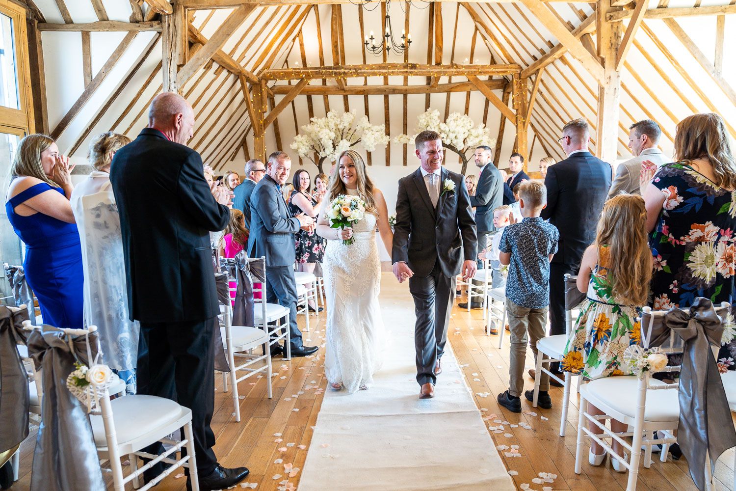 A newlywed couple walks down the aisle in a rustic wooden barn venue surrounded by wedding guests and white chairs.