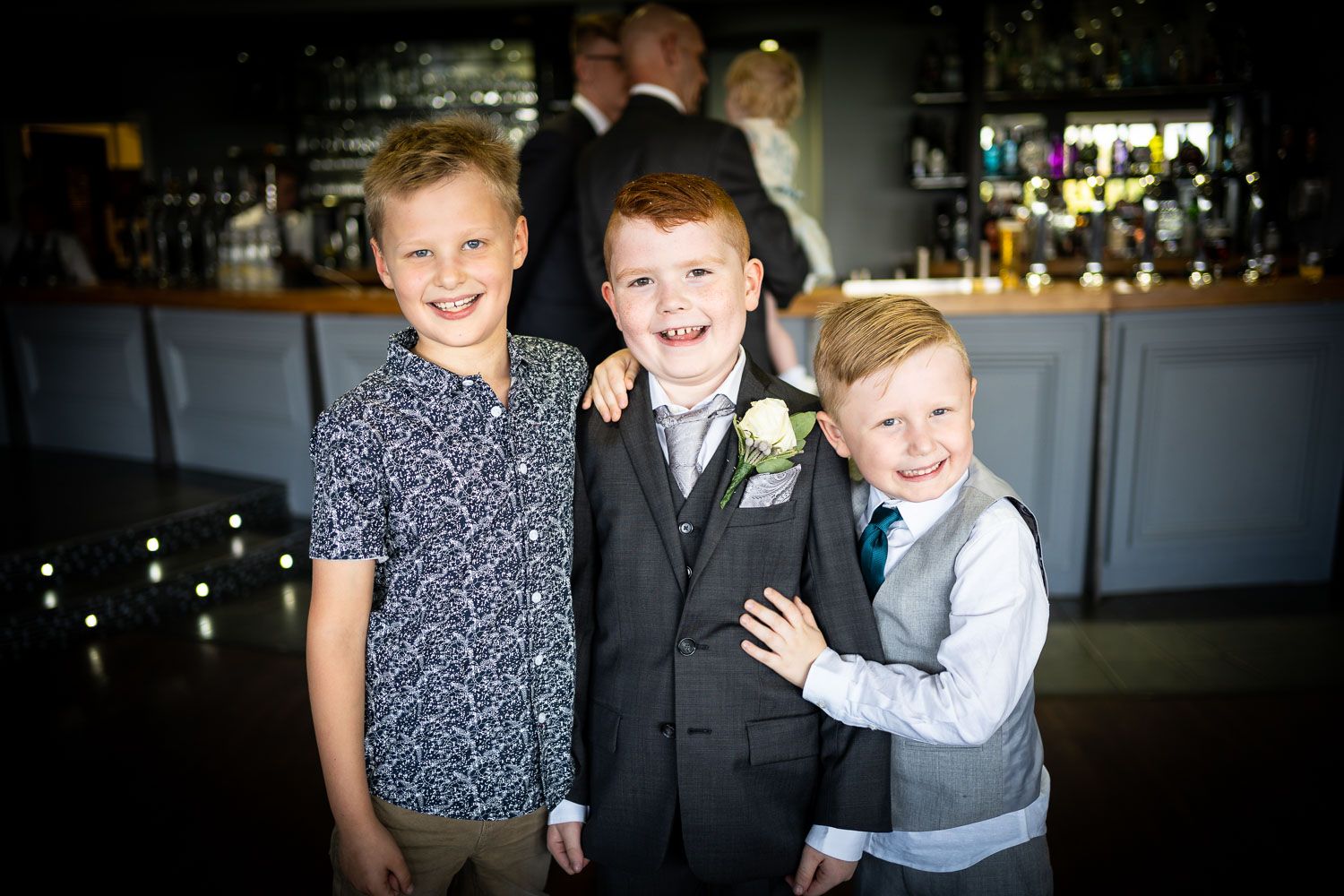 Three young boys dressed in formal and semi-formal attire pose together at an indoor venue with a bar in the background.