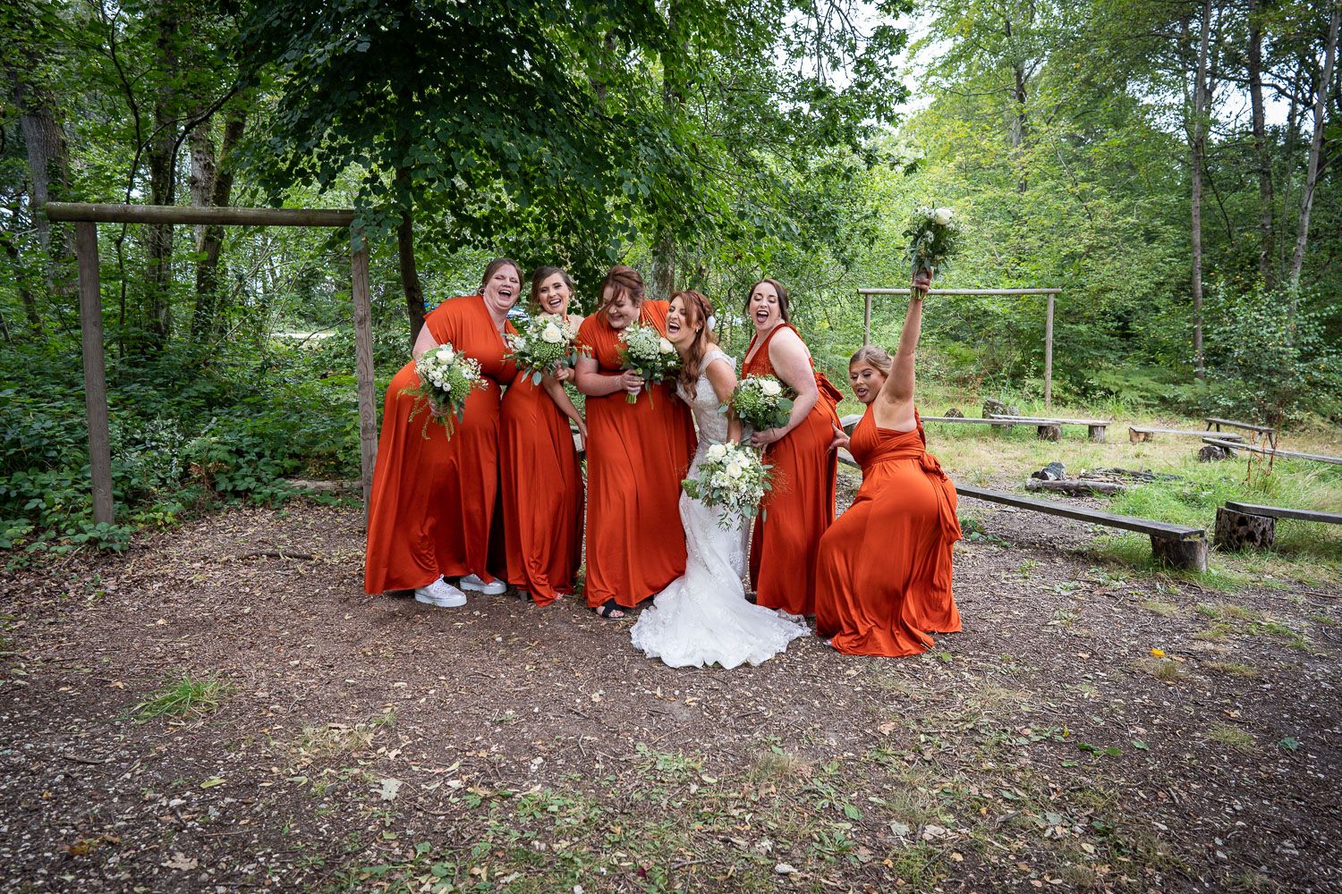 Bridal party in bright red dresses poses together on a wooded path during an outdoor autumn wedding celebration.
