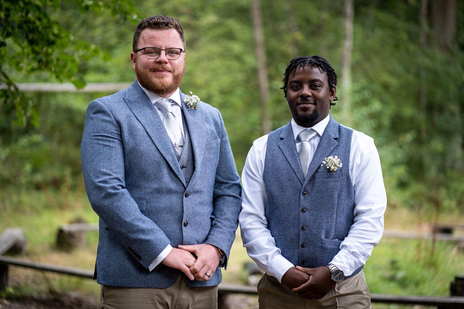 Two groomsmen wearing grey suits pose together on a wooden bridge in front of lush green foliage.