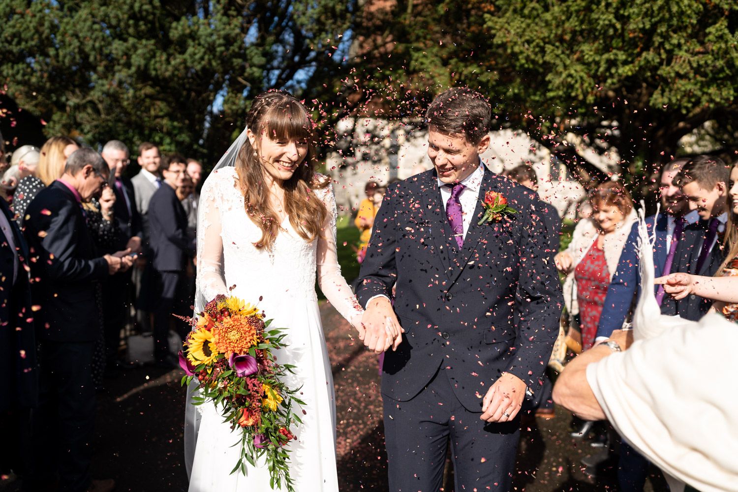 Wedding guests shower newlyweds with confetti outside church on sunny autumn day.