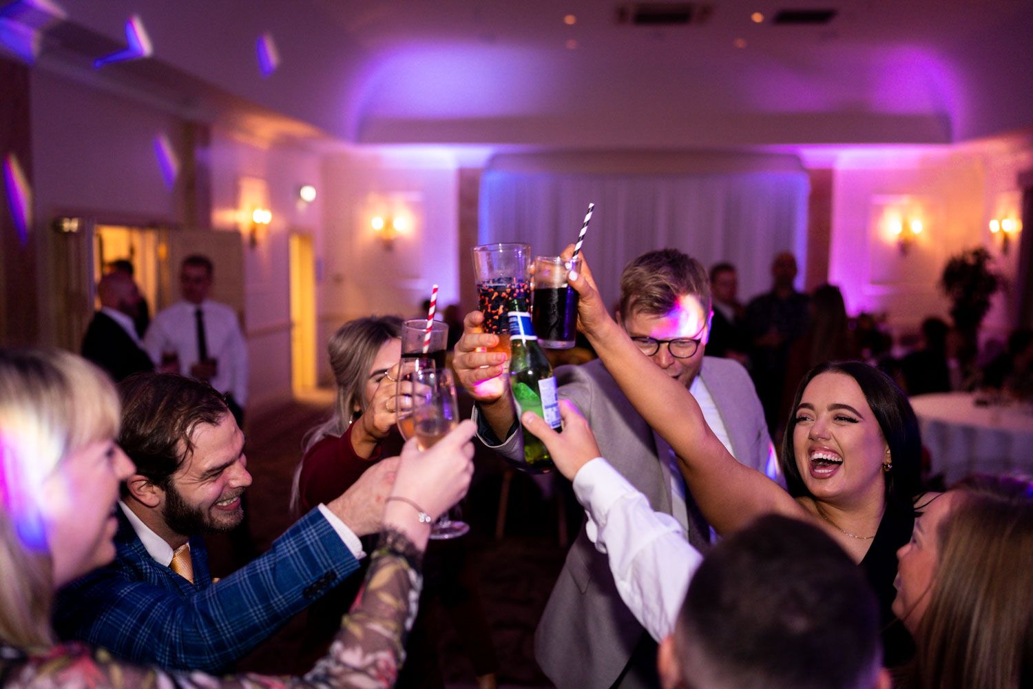 Party guests raise their glasses in celebration with joyful expressions under purple mood lighting at an indoor event.