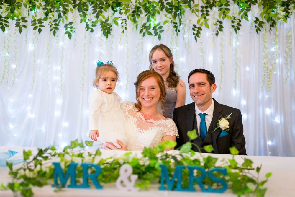 A happy couple sits at a decorated wedding reception table with lighted backdrop and green foliage decor.