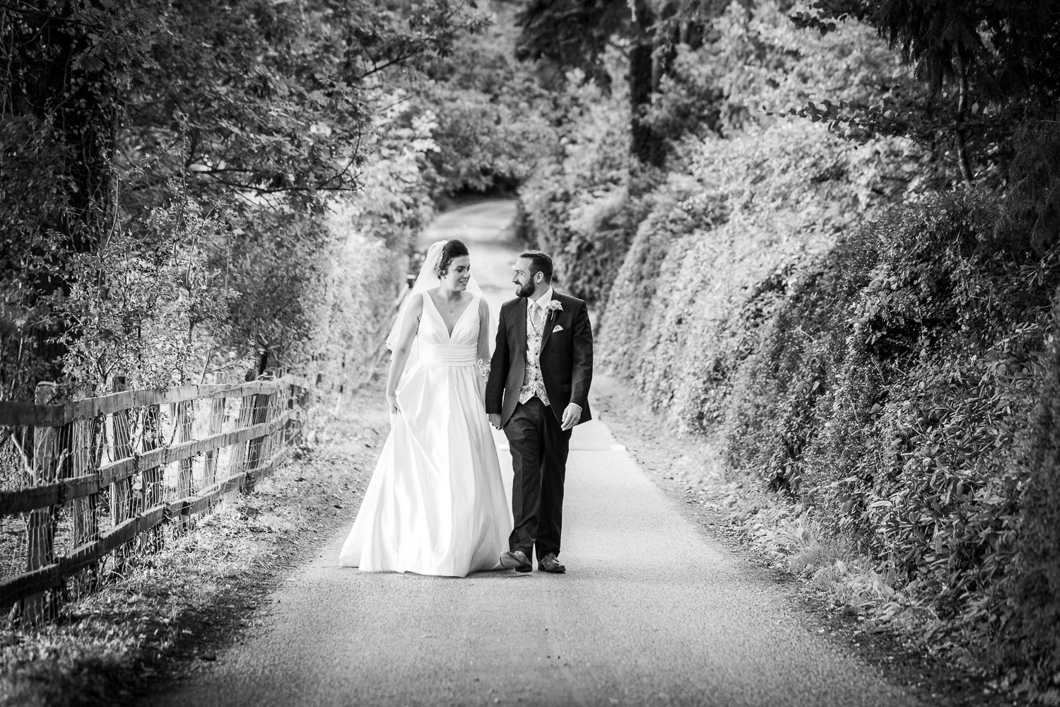 Black and white wedding photo of a couple walking down a scenic path bordered by lush foliage and a wooden fence.