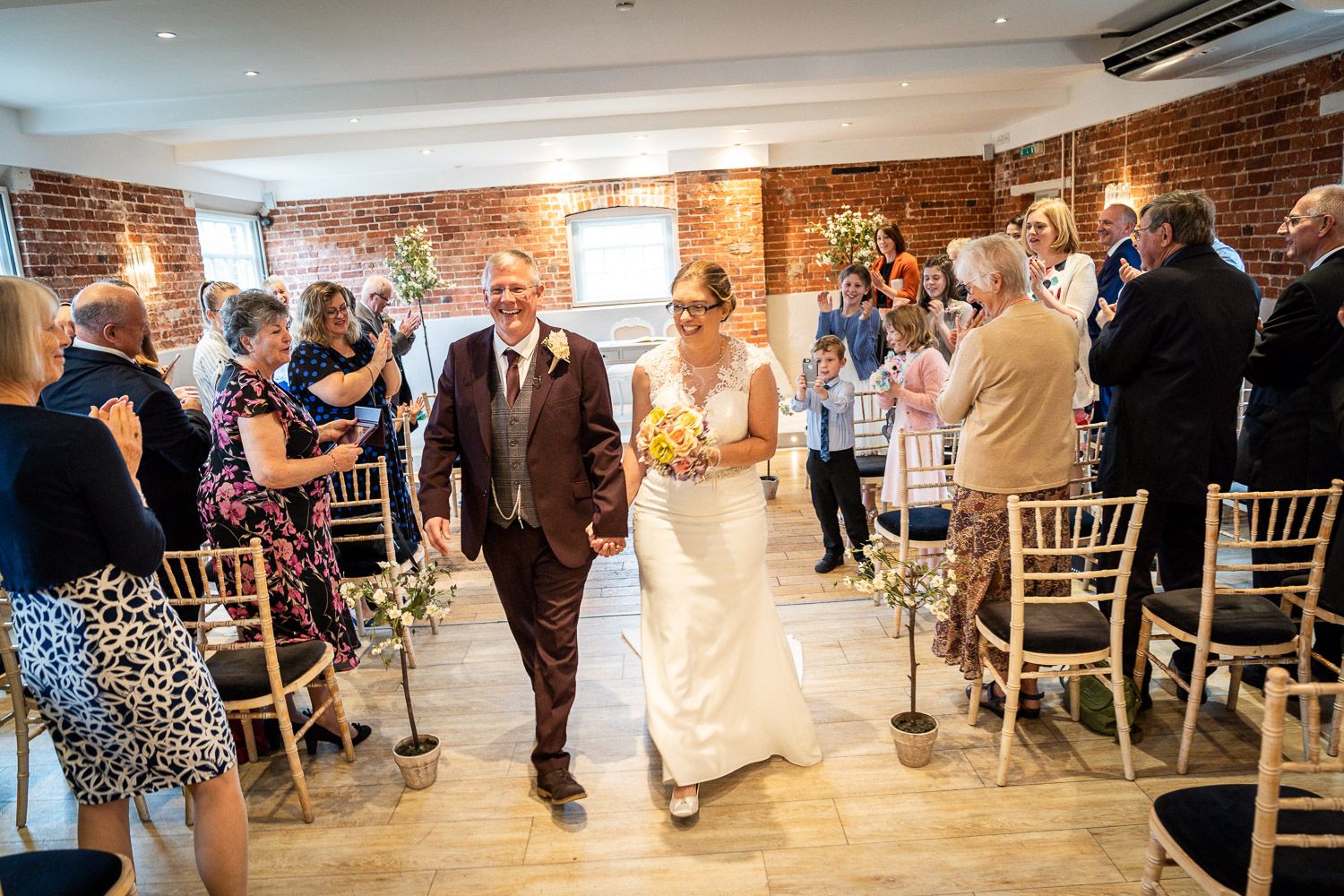 Newlyweds walk down the aisle together in rustic brick-walled venue with wooden floors and chiavari chairs.