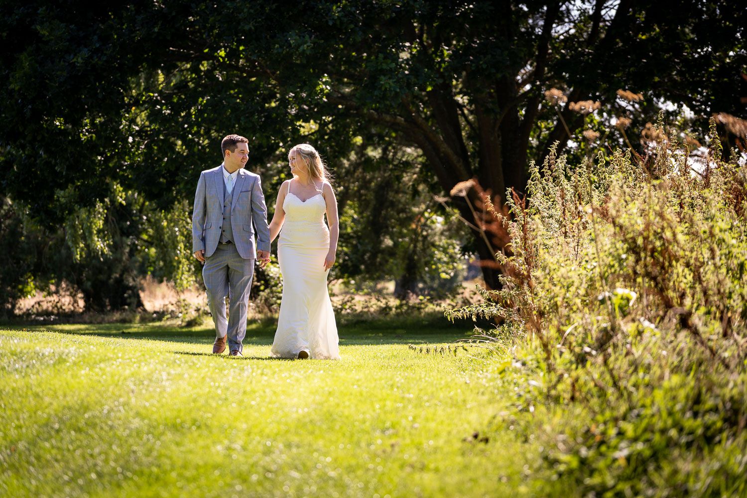 A newlywed couple walks hand in hand through a sunlit garden with tall grasses and trees in the background.