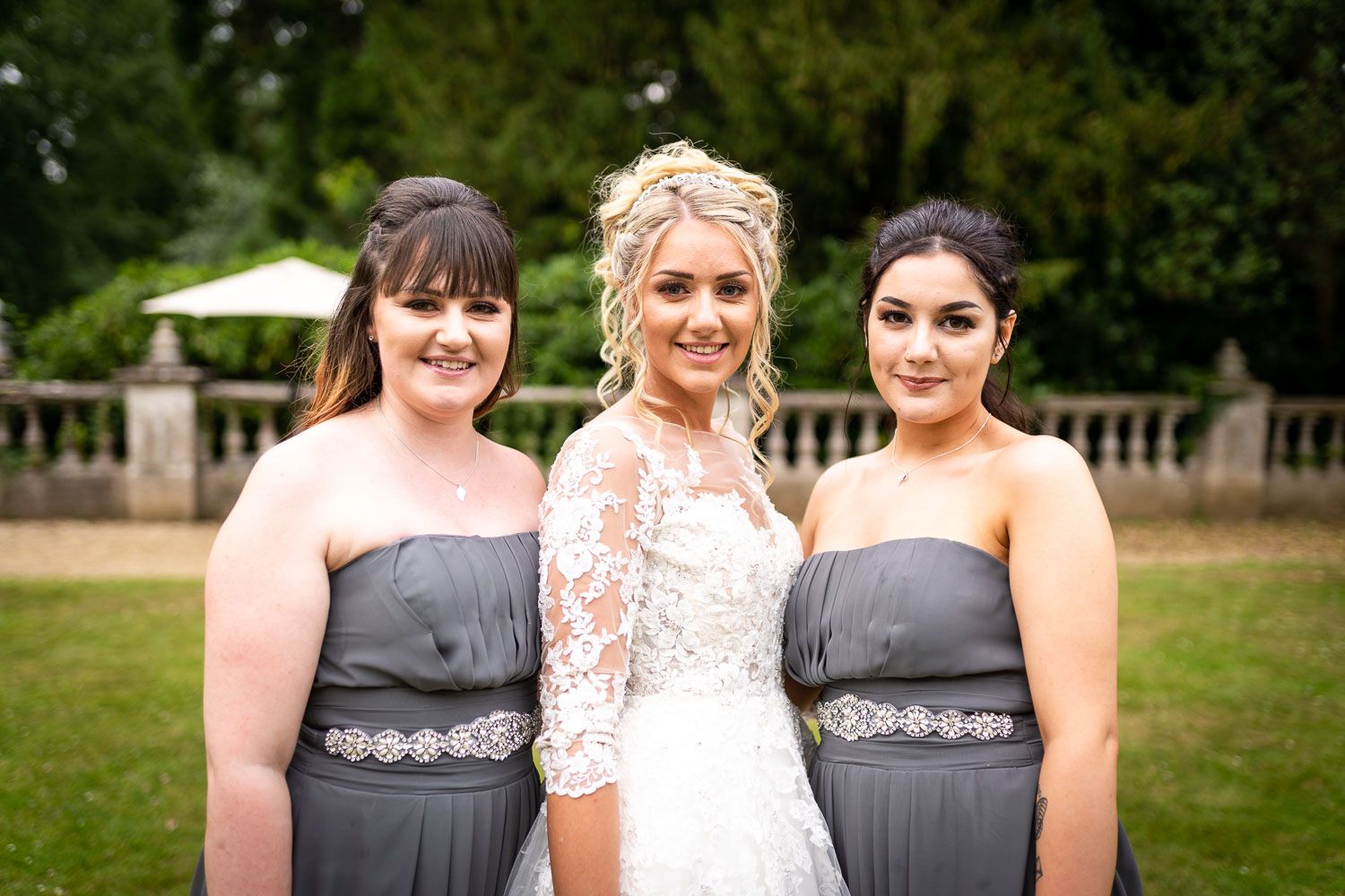 Three women in formal attire pose together outdoors, with one wearing a white wedding dress and two in gray bridesmaid dresses.