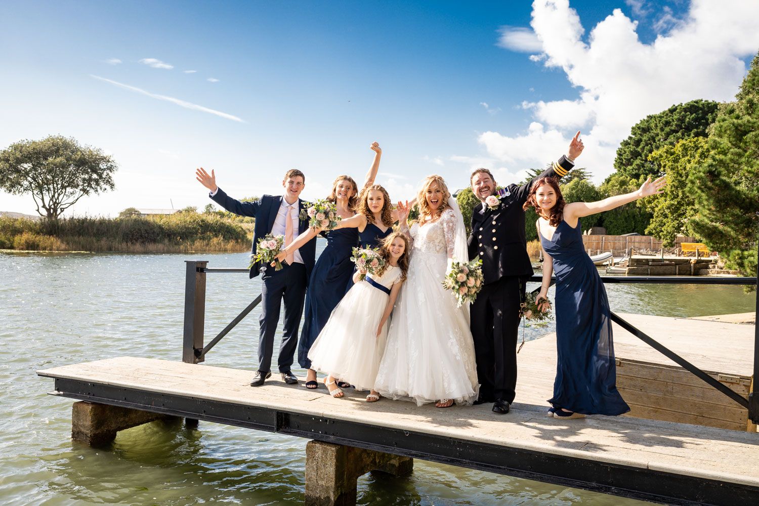 A wedding party celebrates joyfully on a wooden dock by the water on a sunny day with blue skies.