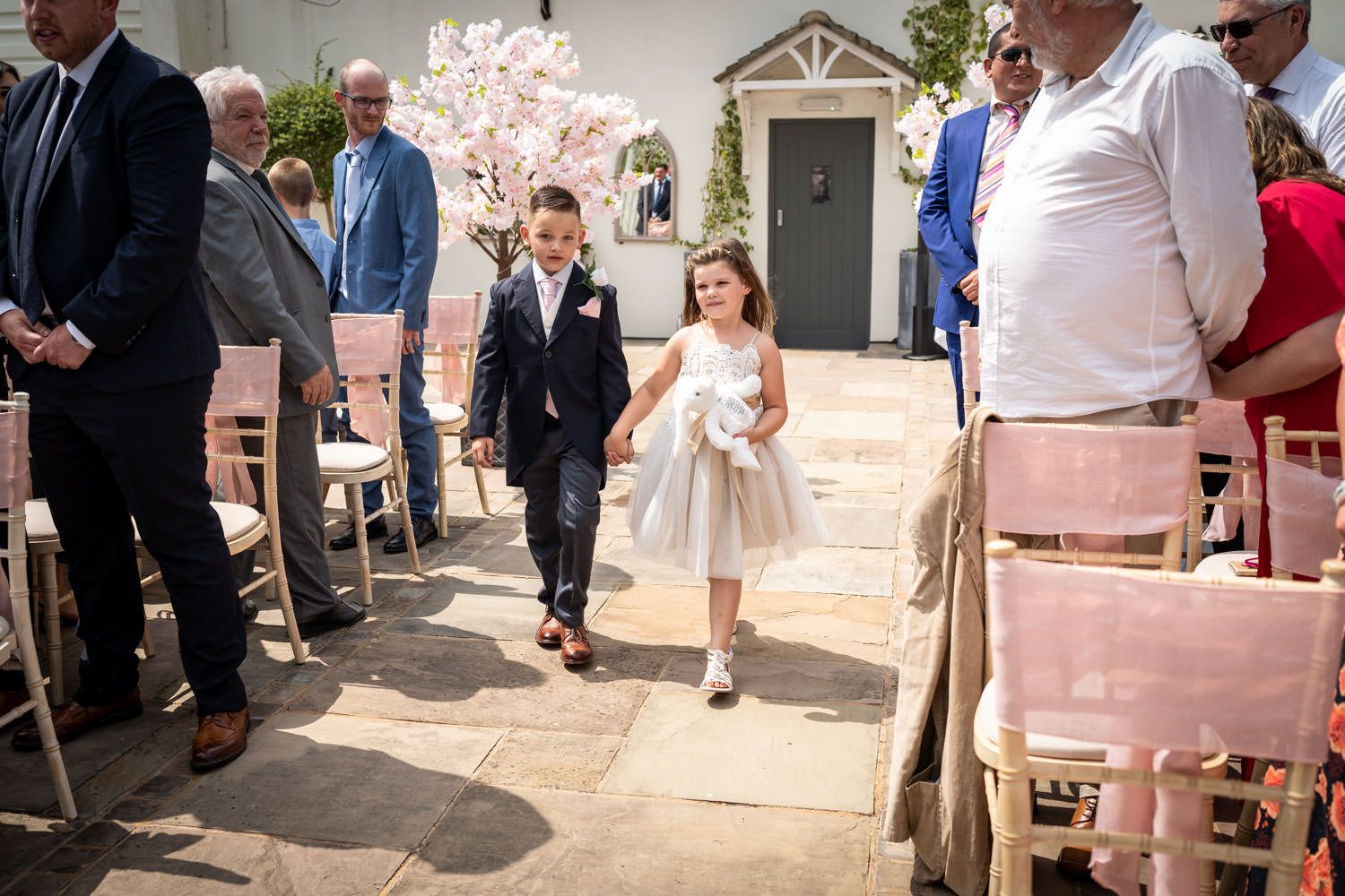 Two young children walk down the aisle together during a wedding ceremony while guests look on from the pews.
