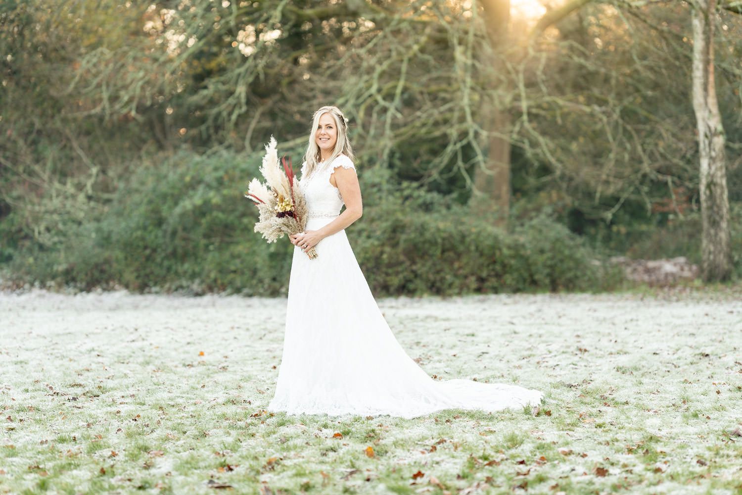 A figure in a white wedding dress holds a bouquet while standing in a sunlit field during golden hour.