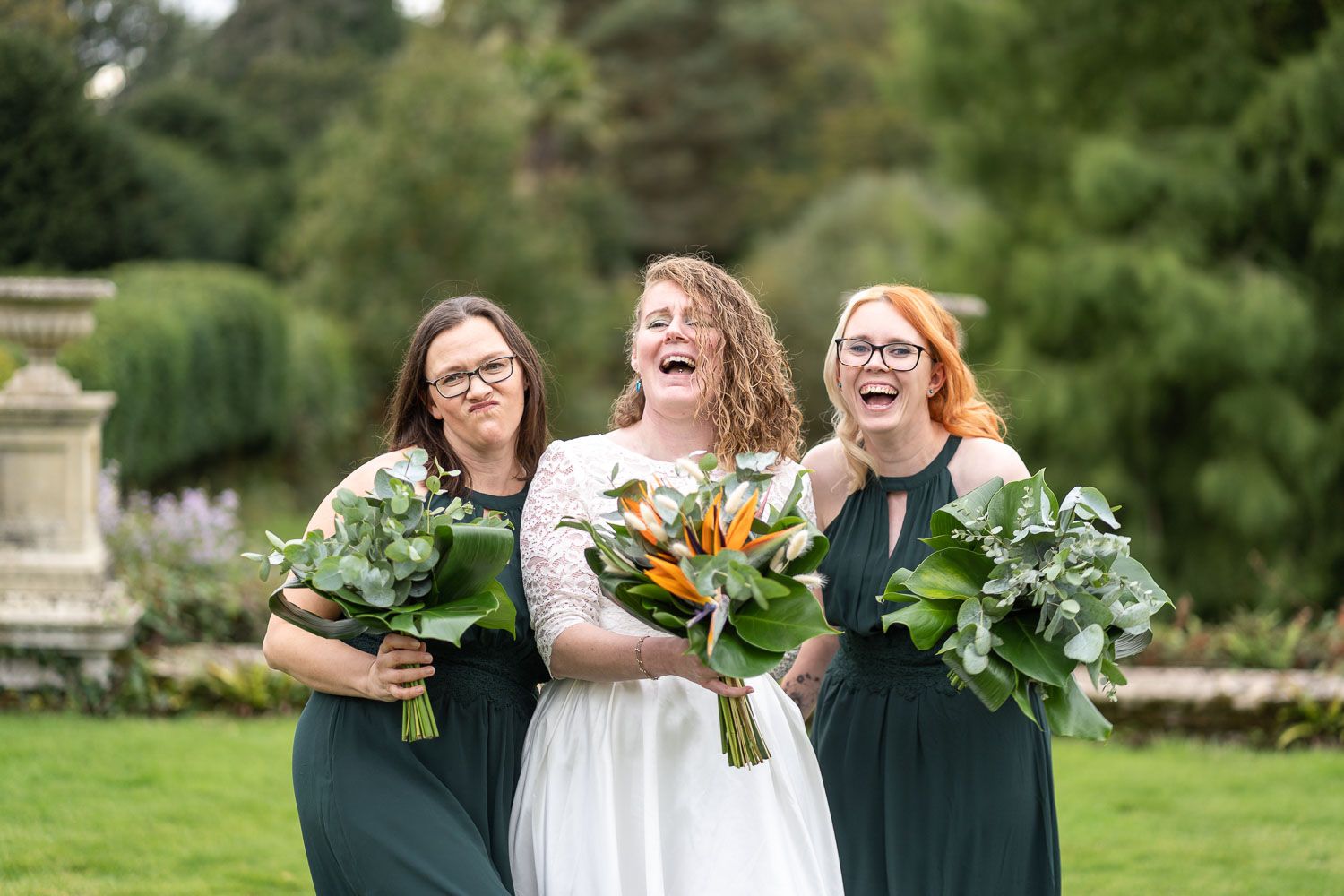 Three women in formal attire sharing a joyful laugh while holding floral bouquets in a garden setting.
