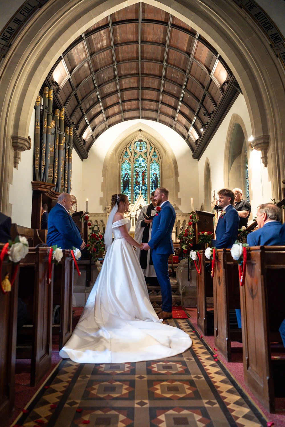 A traditional church wedding ceremony takes place beneath a vaulted ceiling and stained glass window in an ornate sanctuary.