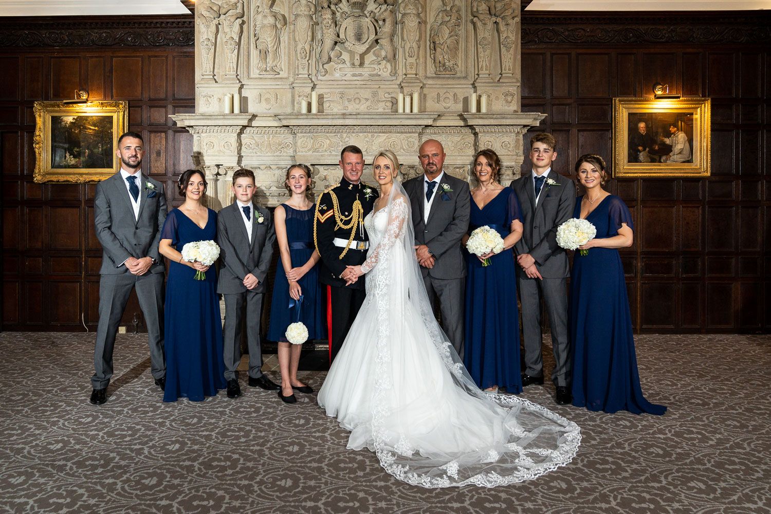 Wedding party in formal attire poses in an elegant wood-paneled room with navy bridesmaid dresses and long white train.