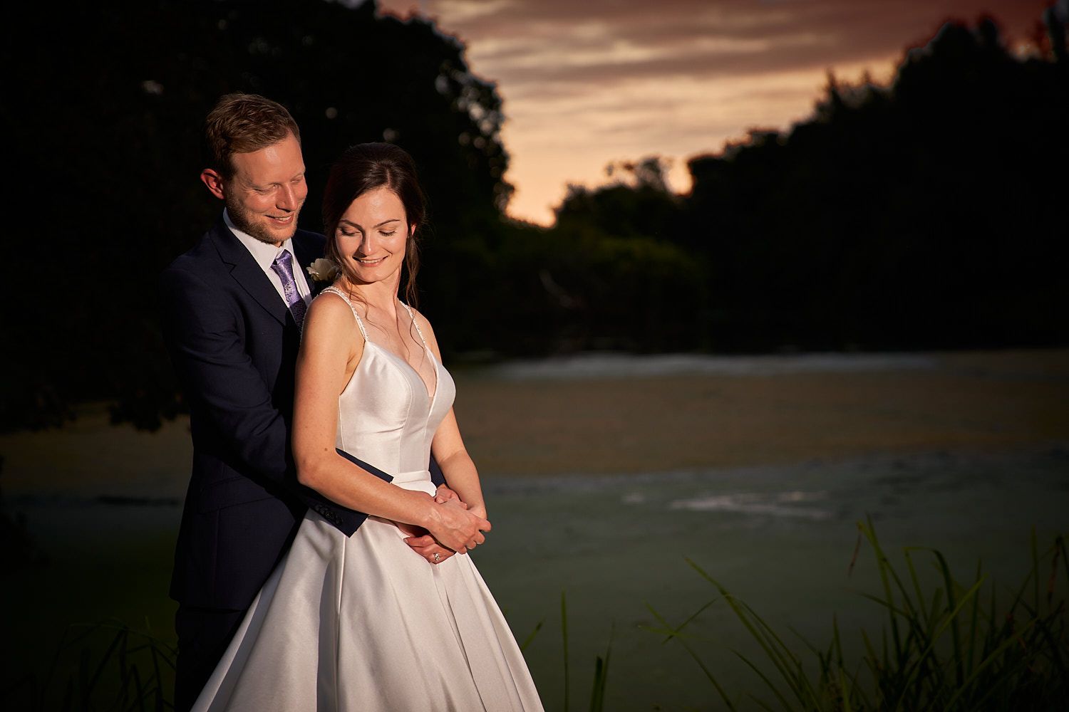 Newlyweds share an intimate moment at sunset by a lake, with soft lighting creating a romantic atmosphere in their formal attire.