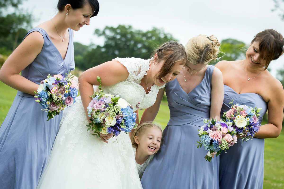A bridal party in powder blue dresses shares a candid laughing moment while holding colorful bouquets outdoors.