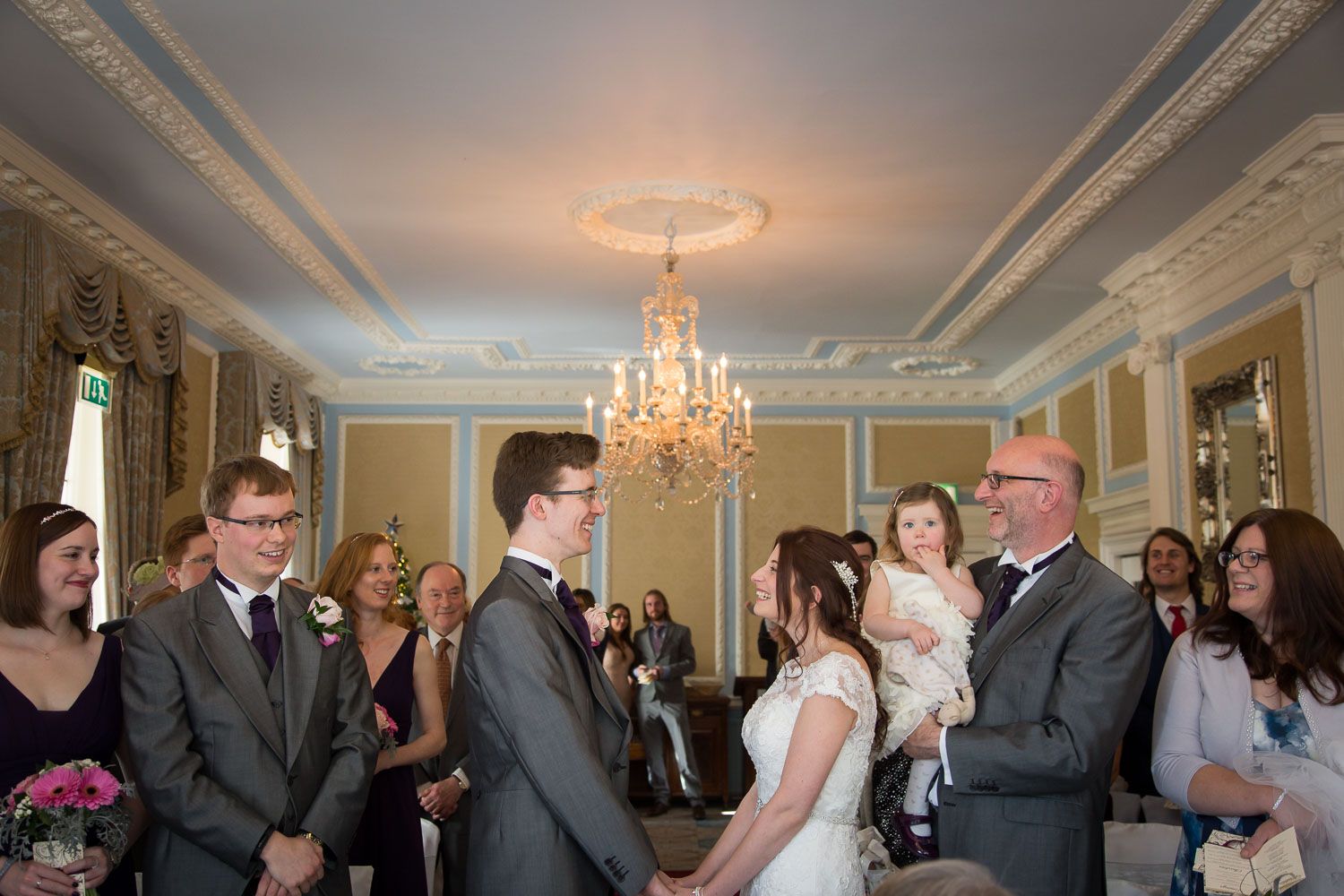 An elegant indoor wedding ceremony takes place under a crystal chandelier in a historic room with ornate moldings.