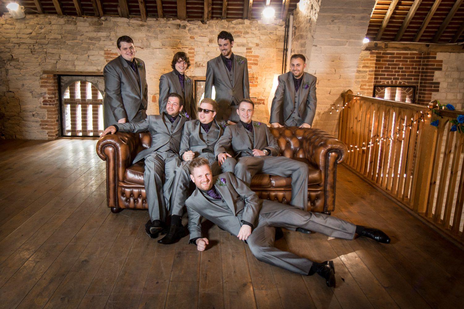 A group in matching gray suits pose together on leather couches in a rustic brick and wood interior space.