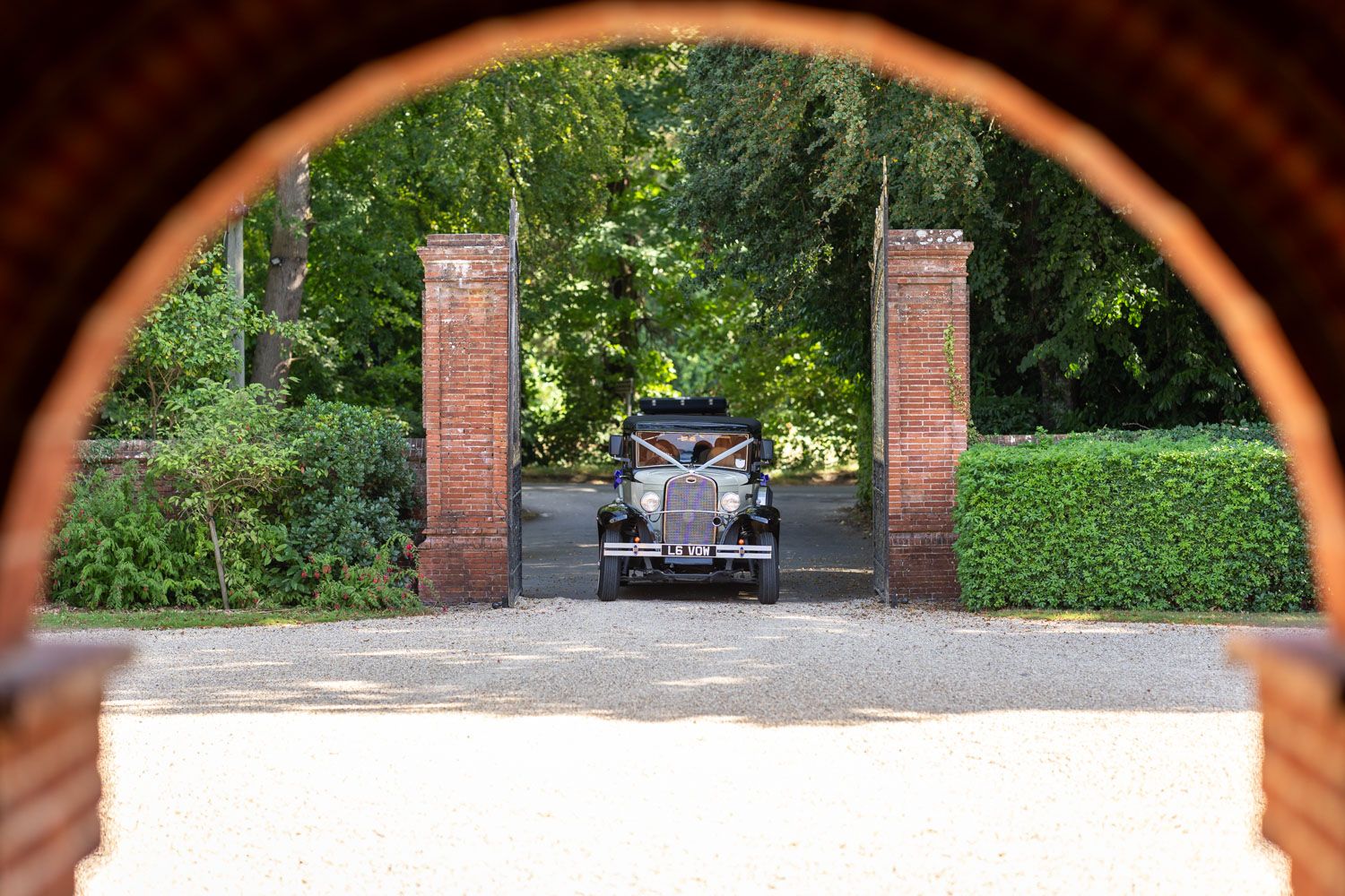 Vintage wedding car arrives through brick gate pillars along tree-lined gravel driveway.