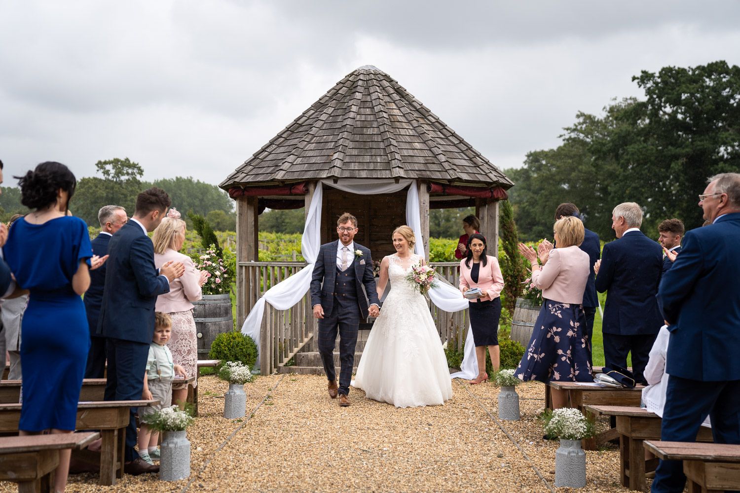 Guests gather at an outdoor wedding ceremony under a wooden gazebo surrounded by lush greenery.