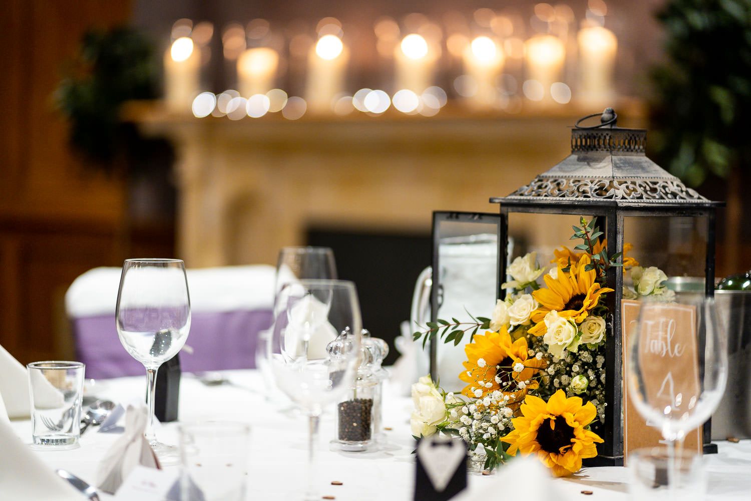 Elegant wedding reception table setting with lantern centerpiece featuring sunflowers and glowing candles in the background.