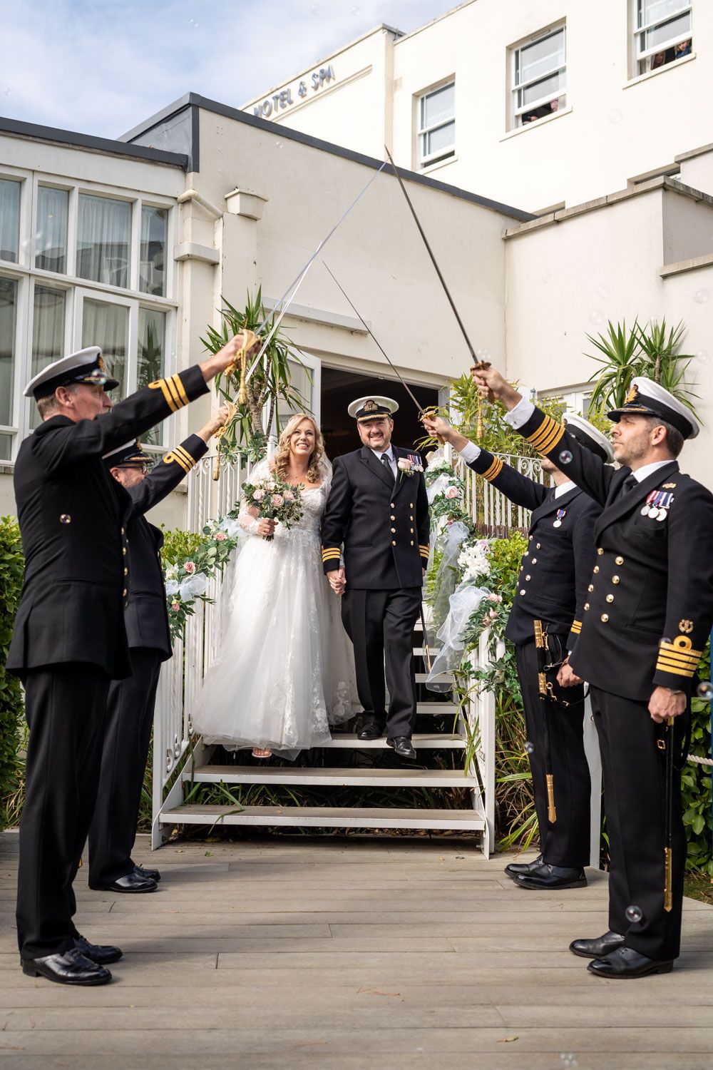 Military officers form an honor guard with raised swords as newlyweds exit down white steps at a formal ceremony.