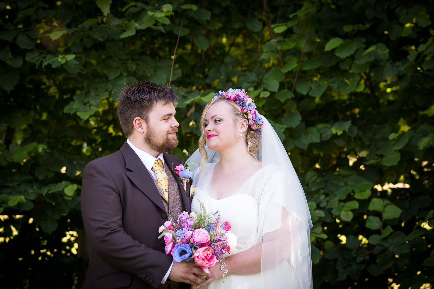 Wedding couple in brown suit and white dress hold pink and purple bouquet against leafy green background.