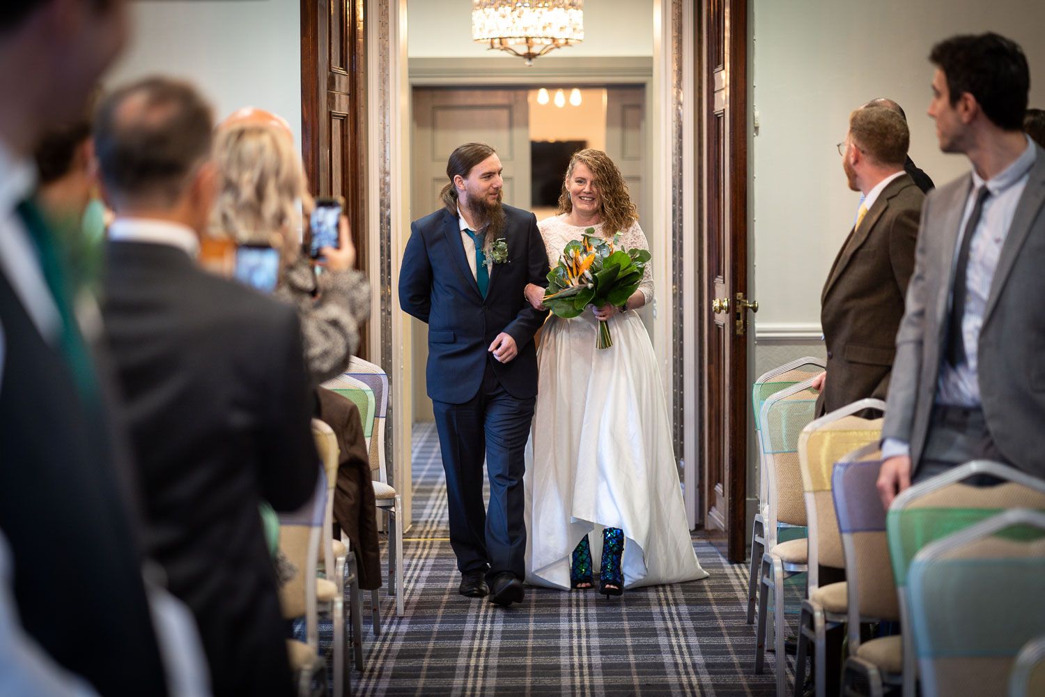 A couple enters a hotel ballroom through double doors while wedding guests watch from both sides of the aisle.