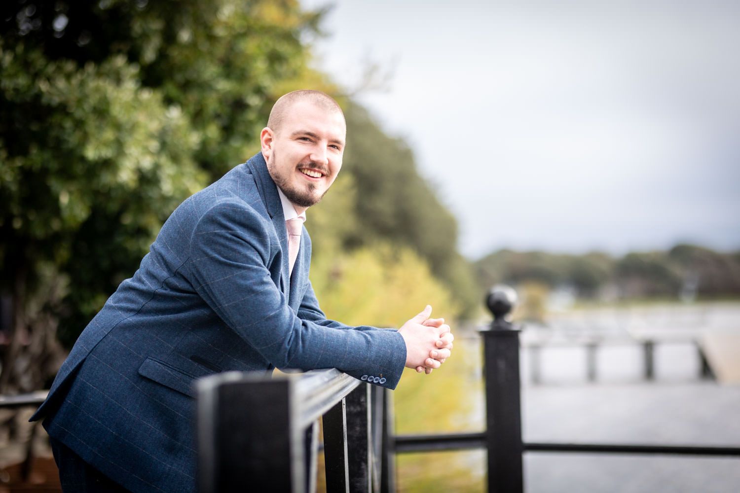 A person in a navy suit leans casually against a black metal railing outdoors with trees and greenery in the background.