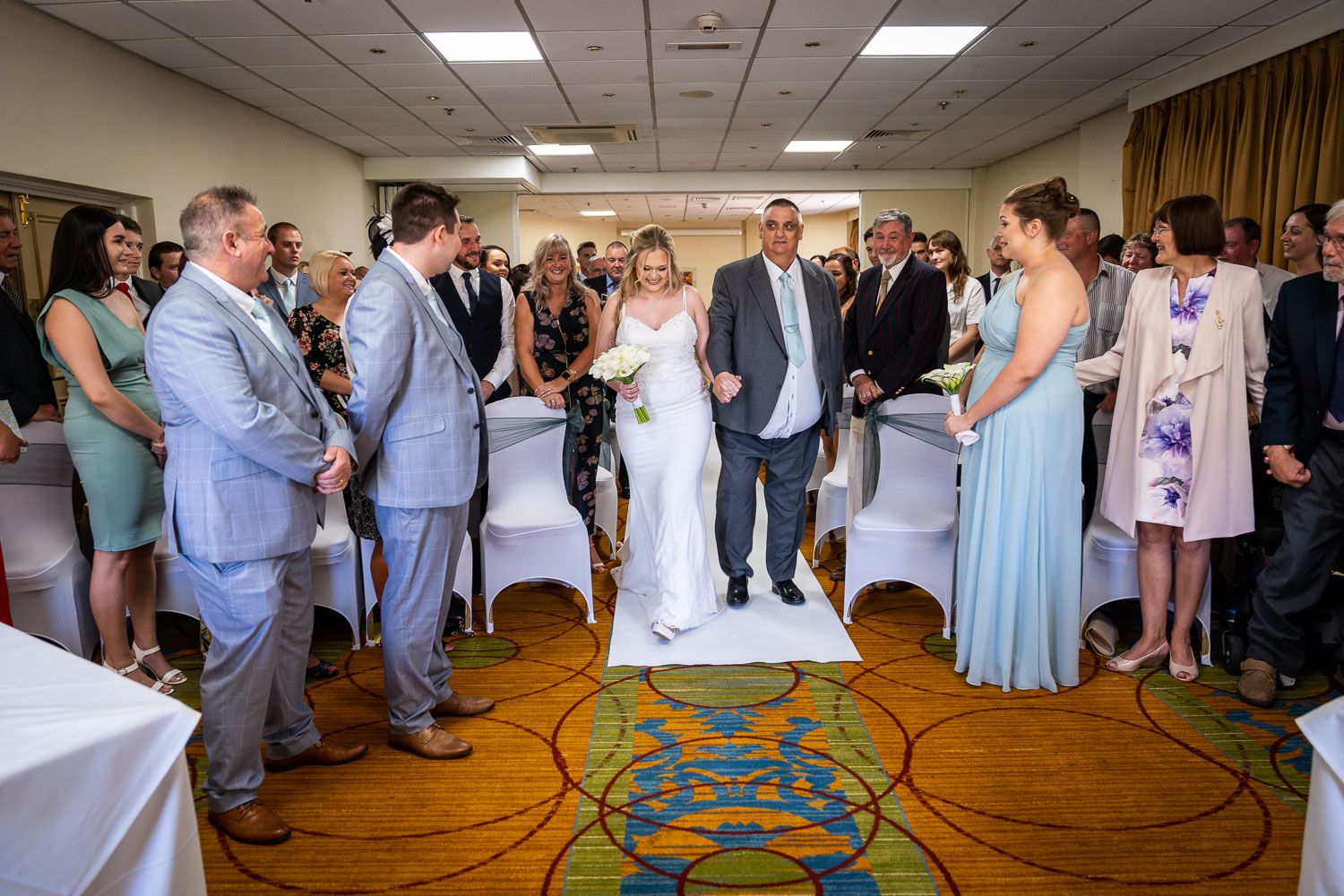 Wedding ceremony guests gather in a reception room with light gray and blue decor around a patterned carpet aisle.