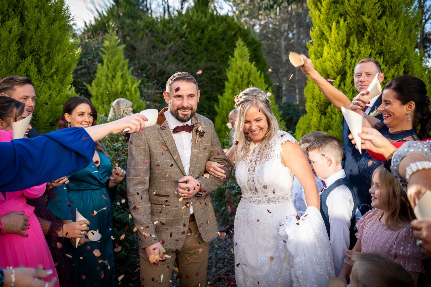 Wedding party celebrates outdoors with confetti while surrounded by evergreen trees.