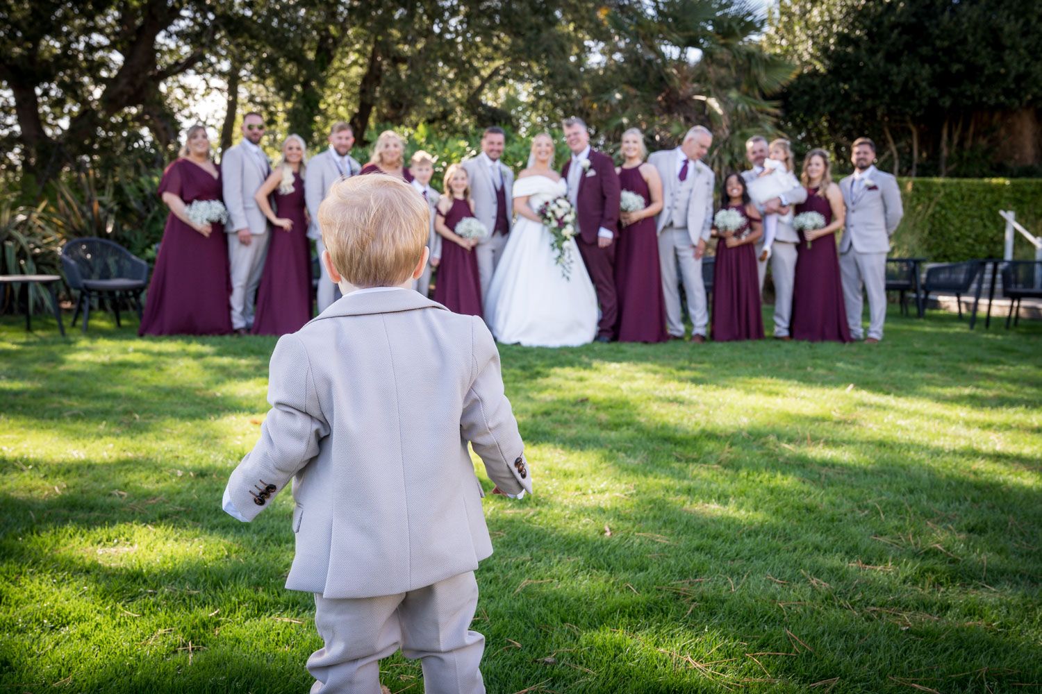 A small child in a gray suit watches a wedding party on a sunny lawn during an outdoor ceremony.
