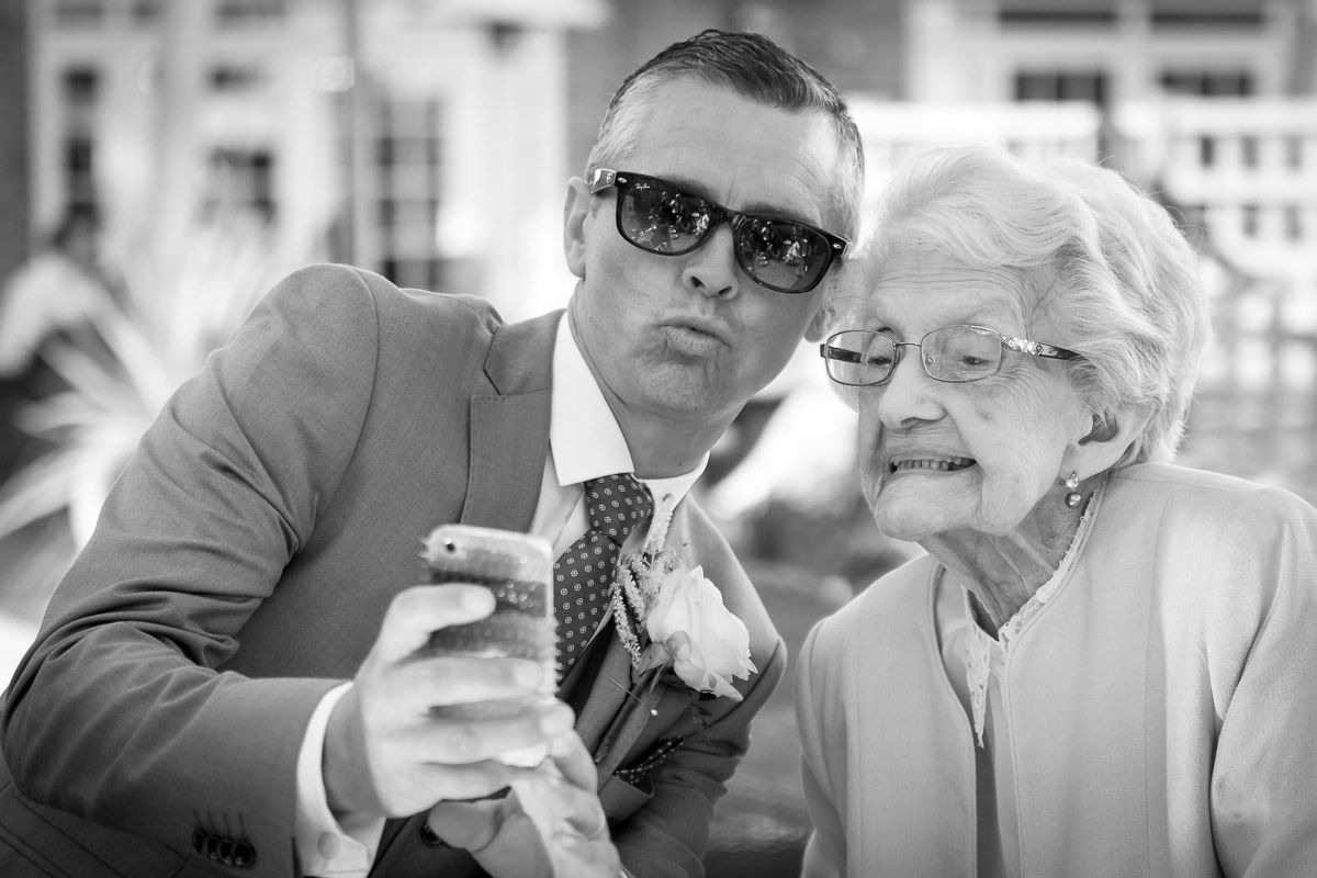 A person in a suit and sunglasses shows something on a mobile phone to an elderly person wearing white.