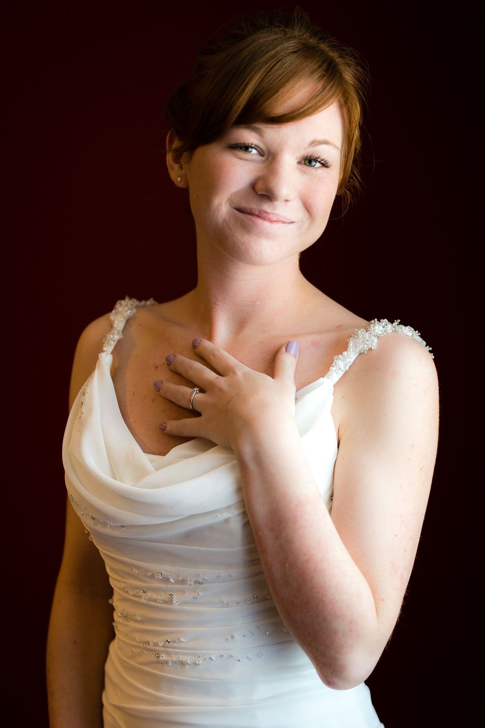 A bride in a white wedding dress with spaghetti straps poses against a dark background with her hand on her chest.